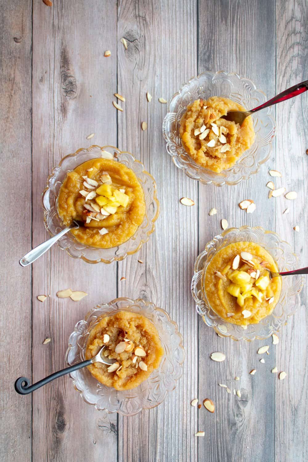 Badam halwa plain and badam halwa with pineapple is served in 4 bowls altogether with spoons in each bowl and sliced almonds are scattered on the background surface.