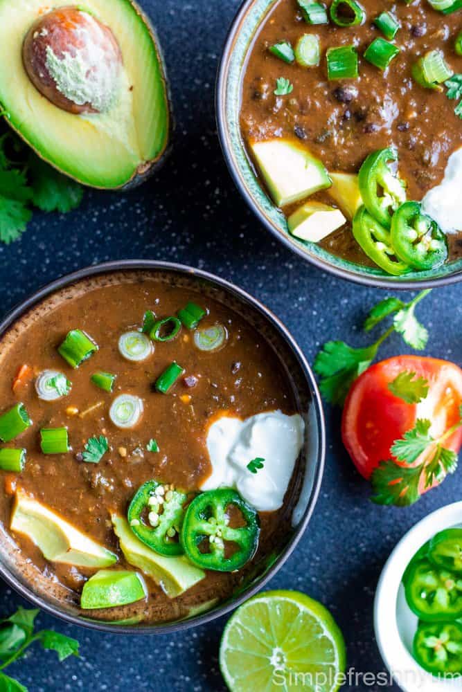 Black bean soup served in two brown color soup bowls with jalapeno slices, sour cream, avocado slices, green onions and cilantro garnished on top with a lemon slice, avocado half on the side with a white bowl of jalapeno slices on the side.