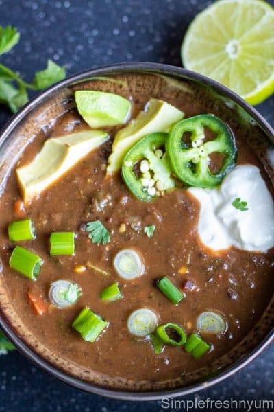 Black bean soup ready to eat in a brown soup bowl with jalapeno slices, sour cream, avocado slices, green onions and cilantro