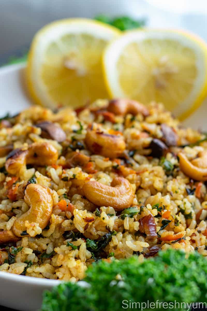 Close up image of Kale rice with cashew nuts garnished on top served on a white plate with lemon slices and fresh kale to the side for of the plate for decoration.