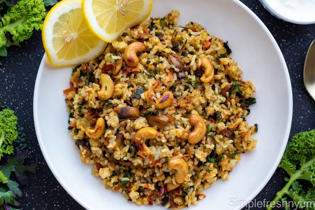 Kale rice with cashew nuts garnished on top served on a white plate with lemon slices. There is a spoon on one side and fresh kale to the side of the plate. Plate is on a black surface with white dots.