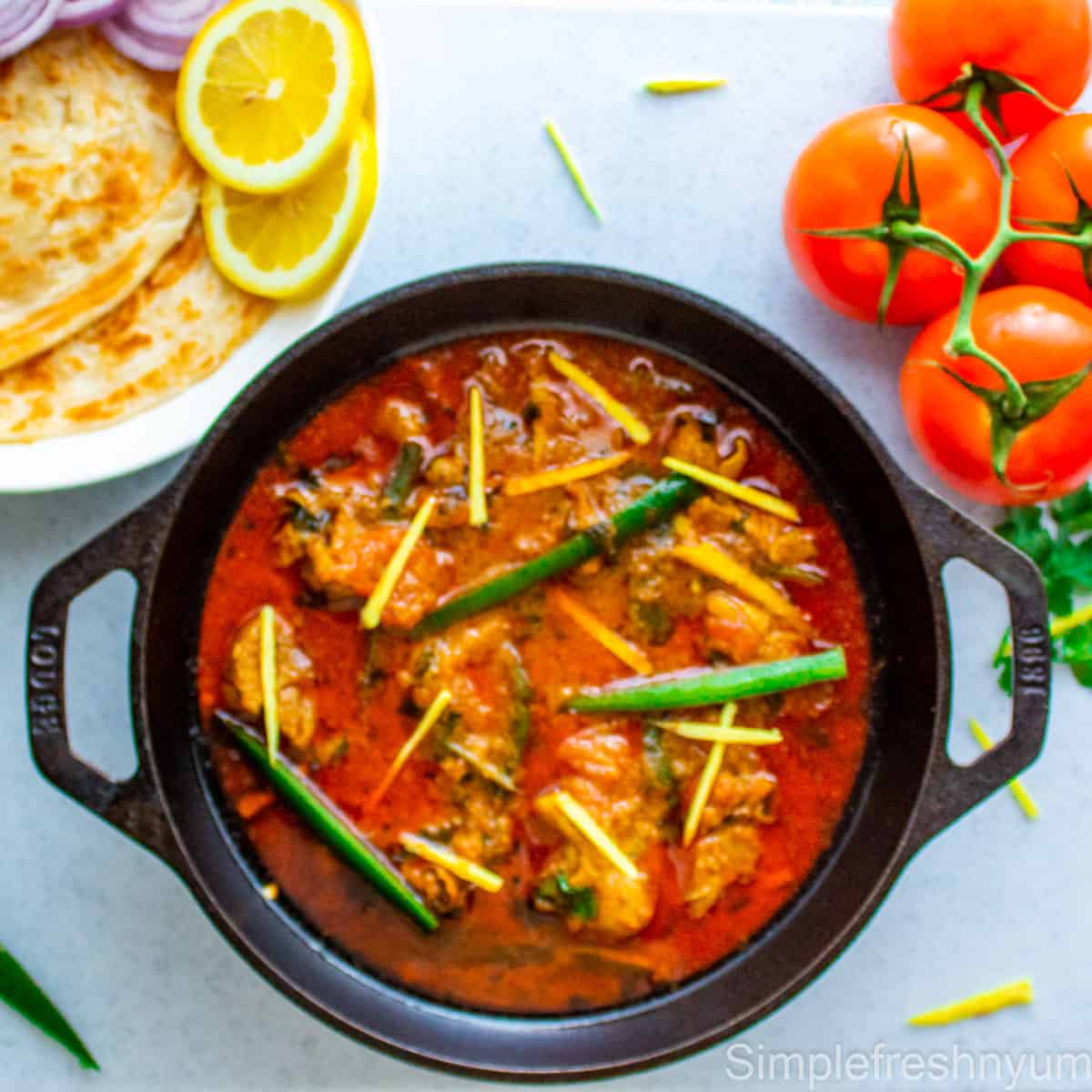 Chicken Karahi in a black cast Iron Wok garnished with thinly sliced jalapenos and ginger juliennes. There are some vine tomatoes on the side and a white plate of paratha with lemon slices on the other side.