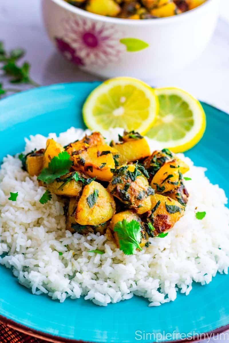 A blue plate with white rice and Aloo palak curry on top with lemon slices on the side of the rice. There is a serving dish with curry in the back ground.