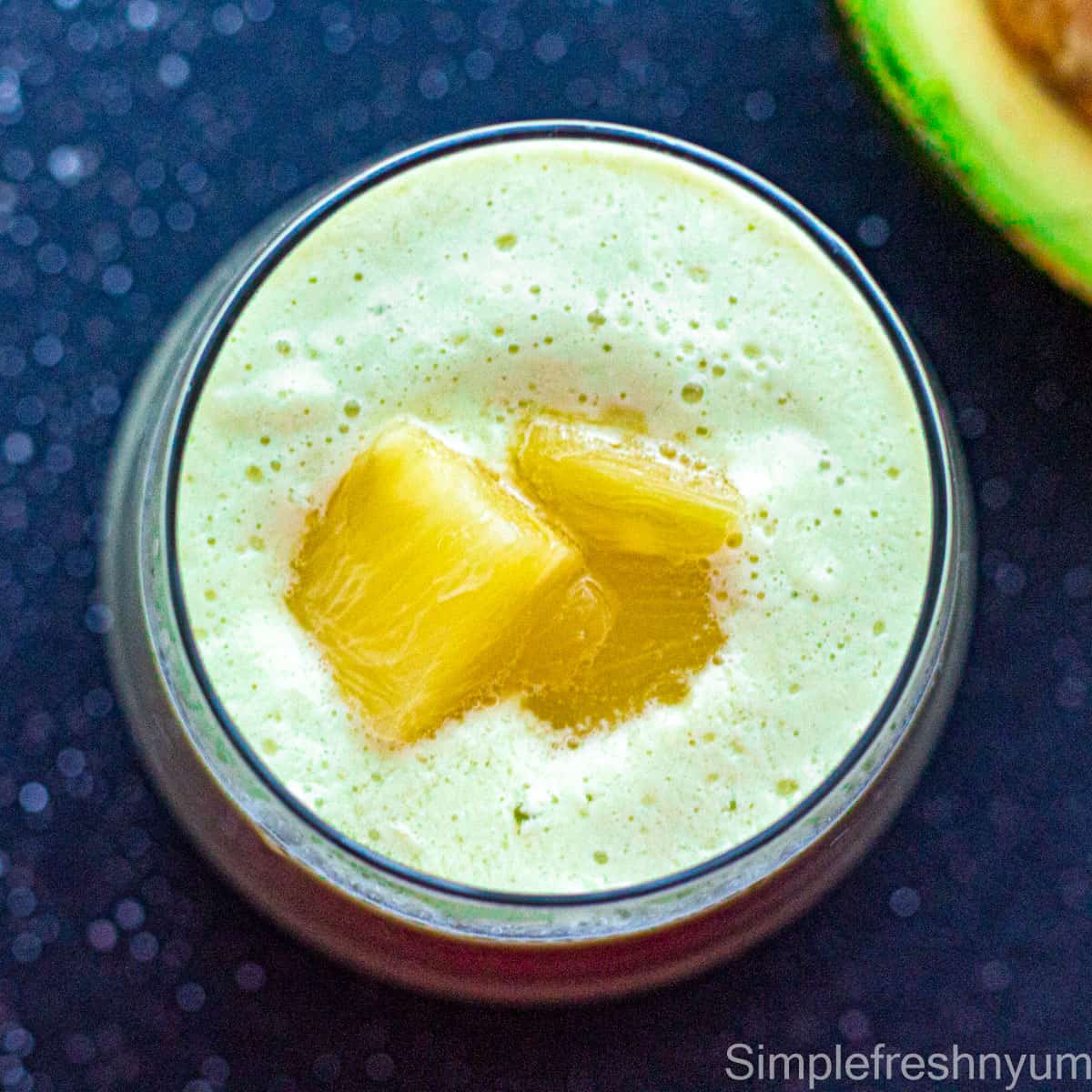 Top view of Avocado pineapple smoothie in a glass with pineapple pieces garnished on top and a piece of half avocado on the side on a black surface.