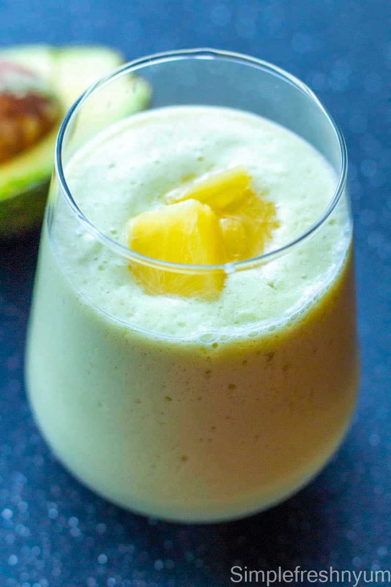 Avocado pineapple smoothie in a glass with pineapple pieces garnished on top and a piece of half avocado on the side on a black surface.
