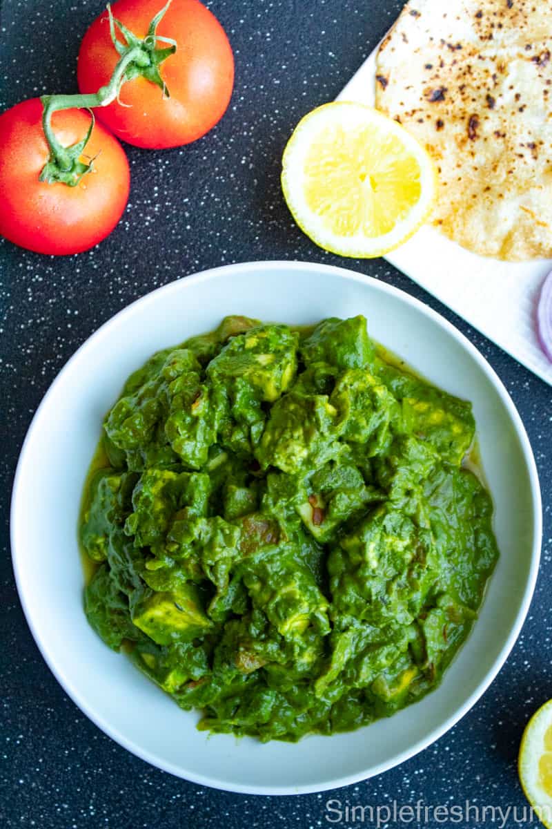 Palak Paneer on a white serving plate with a plate of roti and lemon slice on one side and tomatoes on the other side.