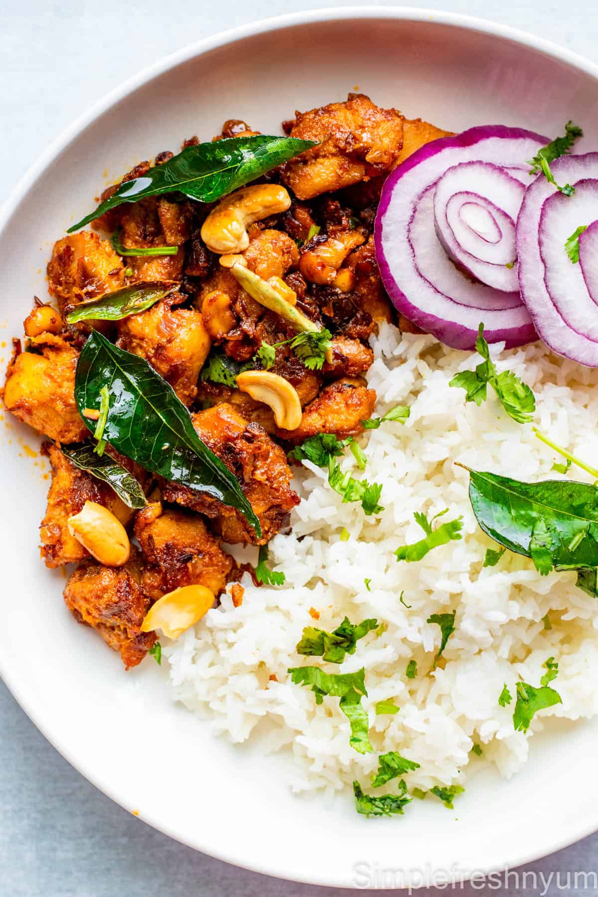 Chicken stir fry served with white rice in a round white plate with sliced onion rounds on the side and cilantro & curry leaves garnished on top.