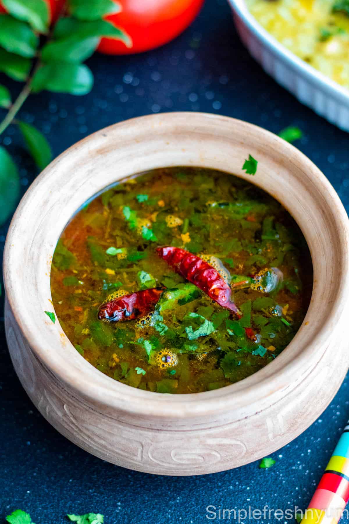 Tomato Rasam served in a clay pot with cilantro and red chilies garnished on top.