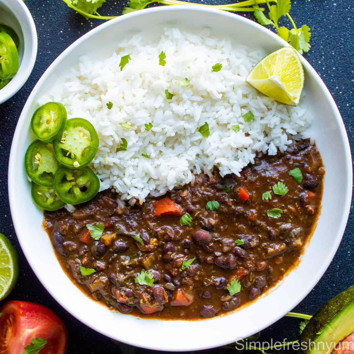 Black bean soup served with rice on a white plate with jalapeno slices and lemon wedge on the side with an avocado half, tomato slice and cilantro on the side of the plate
