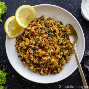 Kale rice served on a white plate with lemon slices on the side and a spoon on the other side on a black surface with fresh Kale to the side of the plate.