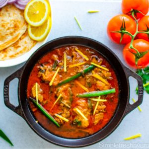 Chicken Karahi in a black cast Iron Wok garnished with thinly sliced jalapenos and ginger juliennes. There are some vine tomatoes on the side and a white plate of paratha with lemon slices on the other side.