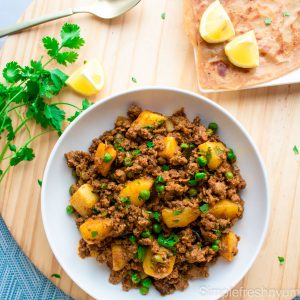 Aloo Kheema Matar served on a white plate with a plate of paratha ( flat bread) and lemon slices on one side and cilantro on the other on a wooden cutting board.
