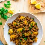 Aloo Keema served in a white plate with a plate of parathas on one side and lemon wedge on the other side on a wooden board.