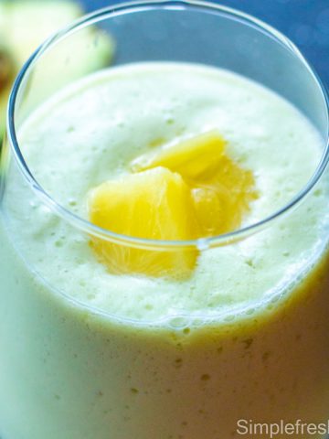 Close up image of Avocado pineapple smoothie in a glass with pineapple pieces garnished on top and a half avocado on the side.