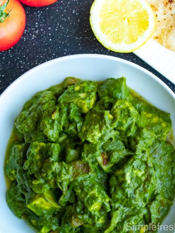 Palak Paneer curry in a white serving plate with red wine tomatoes on one side and a plate of roti with half lemon slice on the other end.