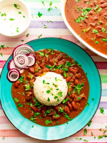 Rajma served in a blue plate with white rice and raw onion rings. There is a bowl of yogurt on the side and a white serving bowl full of Rajma. Cilantro is garnished on top