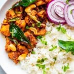 Chicken stir fry served with white rice in a round white plate with sliced onion rounds on the side and cilantro & curry leaves garnished on top.