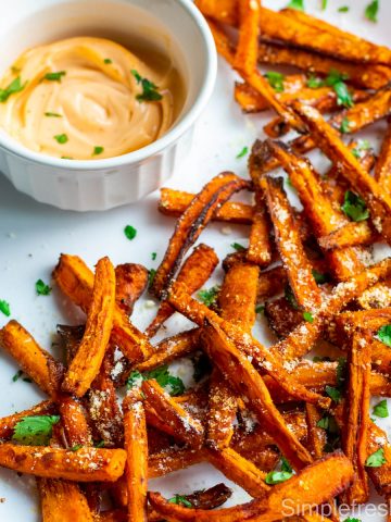 Carrot fries served in a white oval plate with parmesan cheese and cilantro sprinkled on top. Sriracha greek yogurt dip is served in a white round bowl on the side