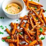 Carrot fries served in a white oval serving plate with parmesan cheese and chopped cilantro sprinkled on top. Sriracha greek yogurt dip is served on the side in a small round white bowl.