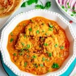 Chicken salan curry served in a white round bowl placed on a bluce plate with cilantro garnished on top. A plate of parotta and another serving dish with sliced onions and lemons are placed on the side.