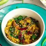 Dondakaya Pachadi served in a white bowl on a round blue plate with tempering on top. Green and red chilies are strewn around the bowl and white rice is served in a bowl in the back ground.
